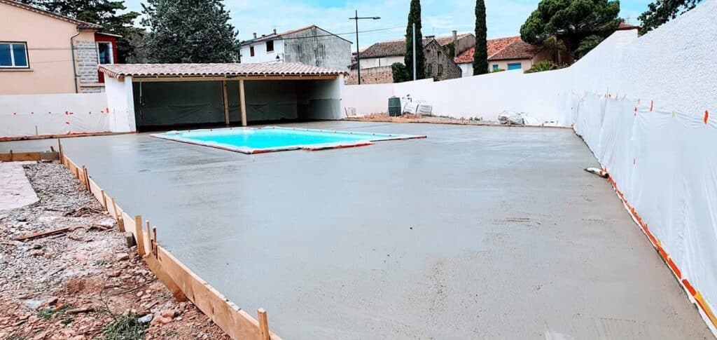 Dalle béton pour terrasse de piscine : étapes de la construction Dalle de béton frais coulée pour une terrasse et une piscine en construction. Coffrage en bois visible devant un mur blanc et un abri.